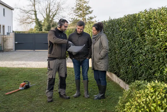 Jardinier équipé échangeant avec des propriétaires devant une haie et une pelouse.