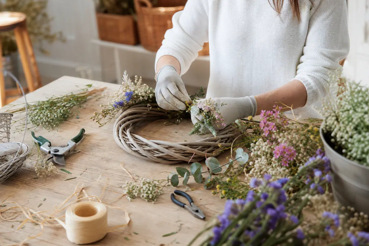 Mains assemblant une couronne de printemps en osier avec fleurs séchées pastel.