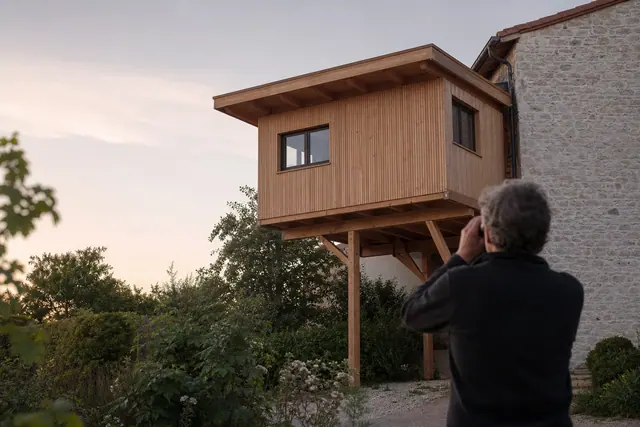 Gîte en bois brut fixé en hauteur sur une façade calme de jardin.