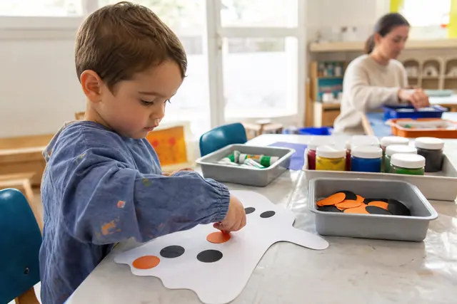 Enfant de maternelle collant de grandes gommettes sur un fantôme en carton.