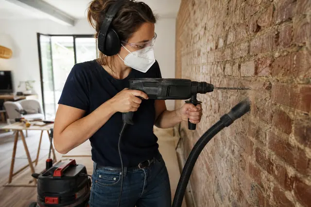 Bricoleuse perçant un mur de brique avec perceuse filaire et lunettes