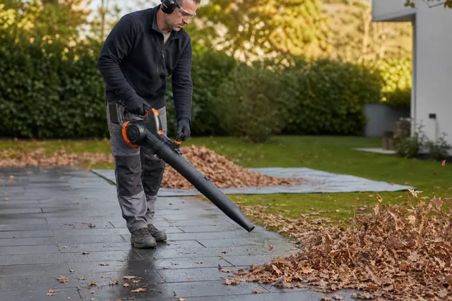 Jardinier protégé utilisant un souffleur sans fil sur une allée couverte de feuilles.
