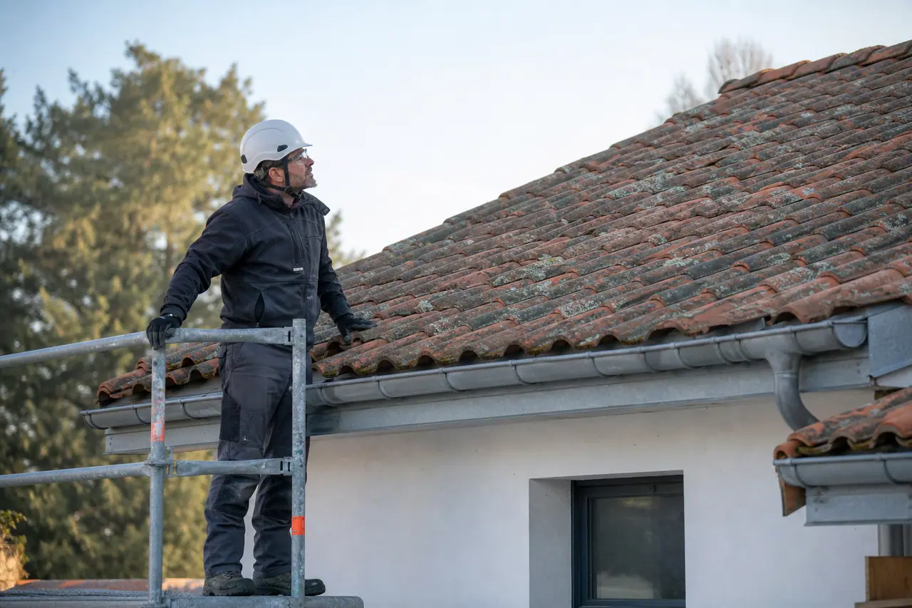 Couvreur inspectant des lichens sur des tuiles depuis un échafaudage sécurisé.