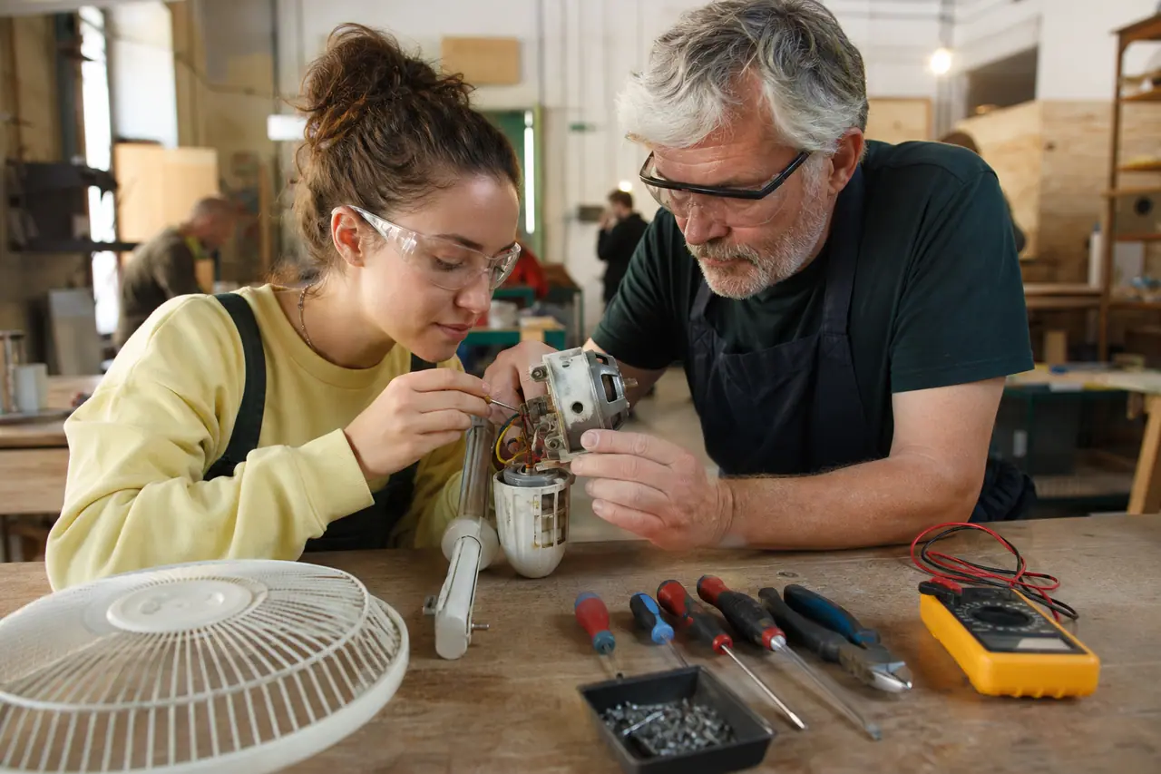 Débutante réparant un petit ventilateur avec un bénévole dans un atelier partagé.