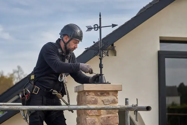 Pose sécurisée d’une girouette métallique sur un pignon maçonné depuis un échafaudage.