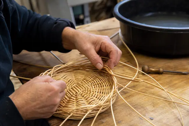 Mains tressant un petit panier rond en rotin sur une table d’atelier.