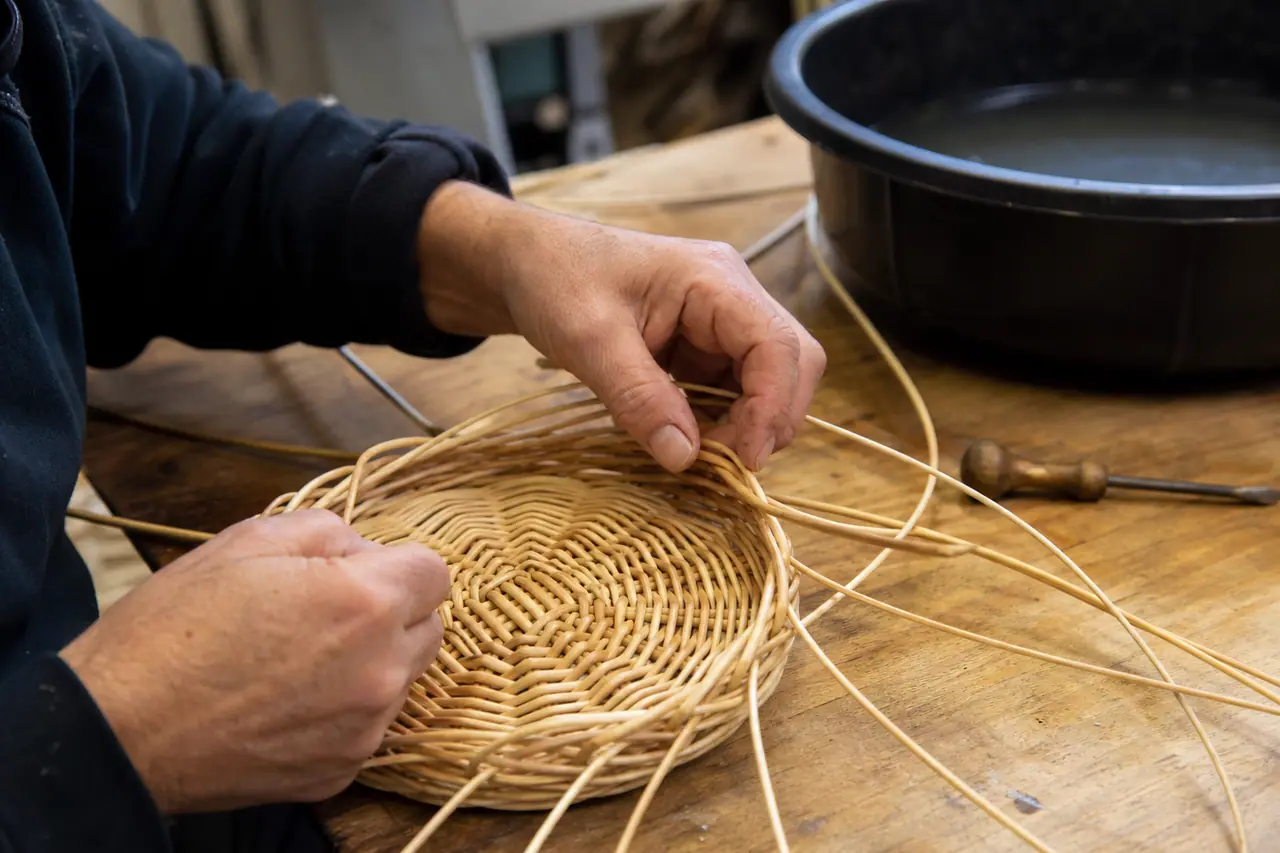 Mains tressant un petit panier rond en rotin sur une table d’atelier.