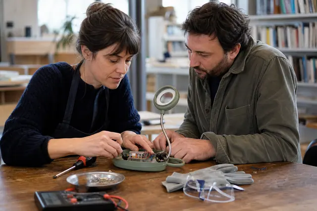 Bénévole et visiteur diagnostiquant une lampe débranchée dans un atelier partagé.