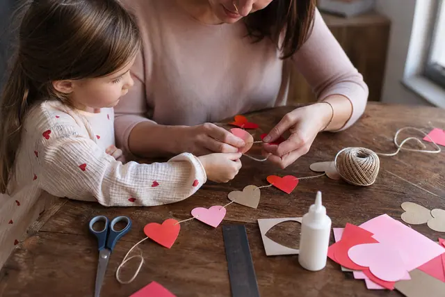 Adulte et enfant fabriquant une guirlande de cœurs en papier sur une table.