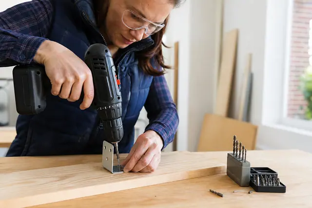 Bricoleuse équipée vissant une équerre sur une planche avec une perceuse sans fil.
