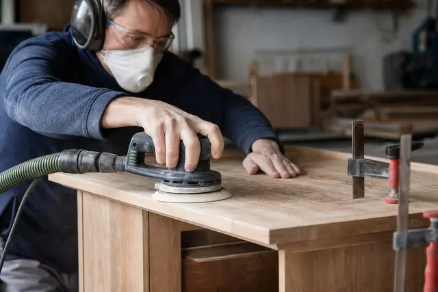 Bricoleur ponçant une commode en bois avec ponceuse excentrique et aspiration.