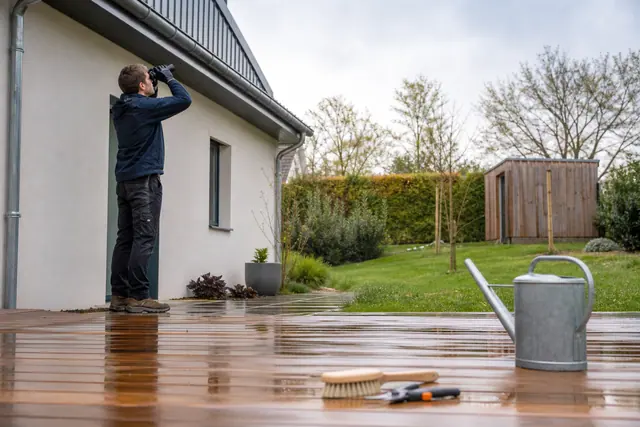 Particulier inspectant une gouttière depuis son jardin au printemps, devant une terrasse en bois.
