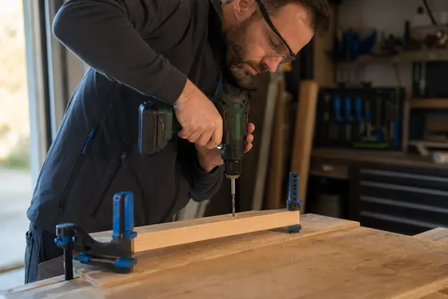 Bricoleur équipé vissant un tasseau sur établi avec une perceuse sans fil.