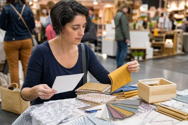 Visiteuse comparant tissus et objets en bois sur un stand créatif à Montpellier.