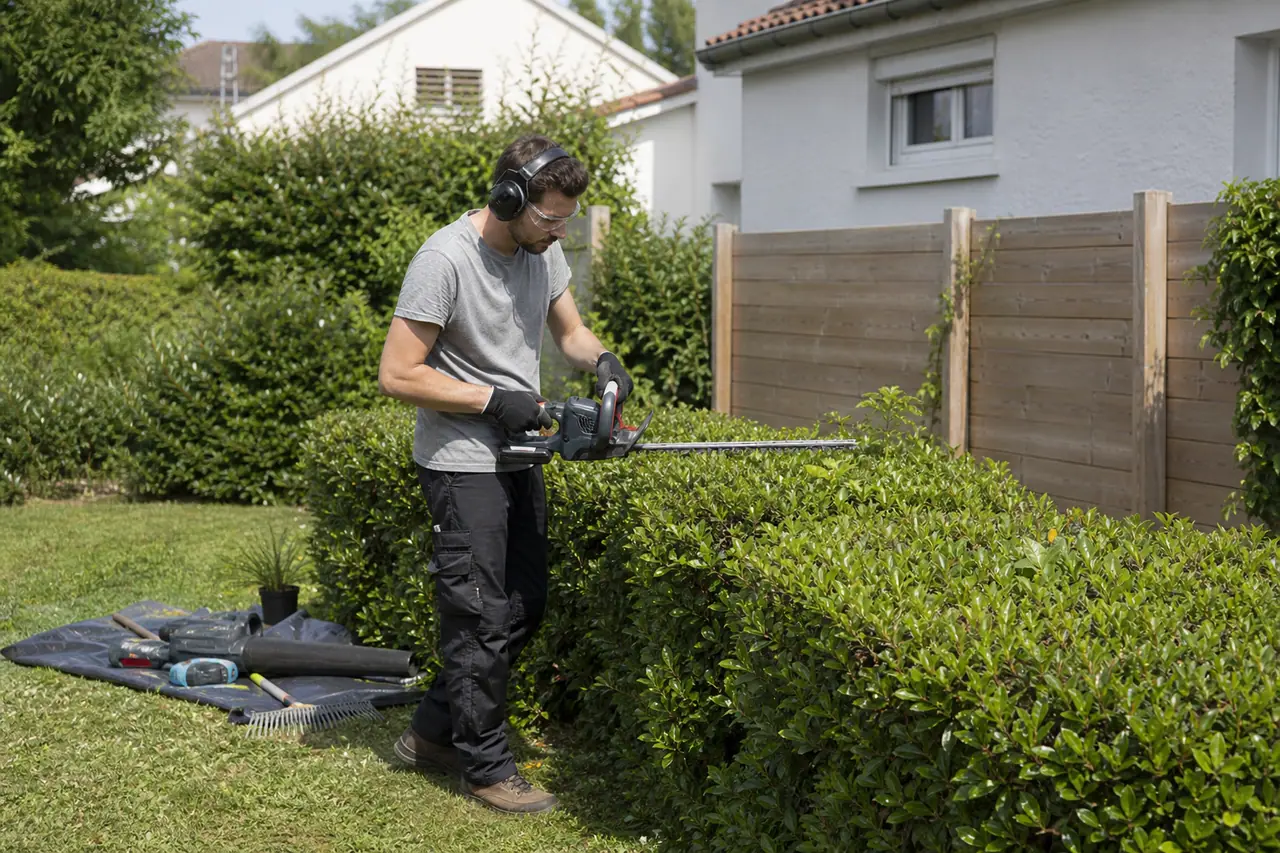 Jardinier équipé utilisant un taille-haie sur batterie dans un jardin résidentiel calme.