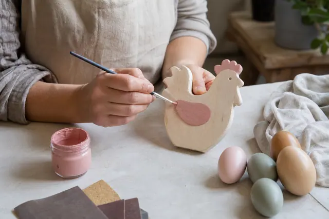 Personne peignant une poule en bois avec des couleurs pastel sur table d’atelier.
