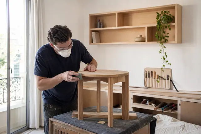 Personne ponçant une table d’appoint en bois dans un salon lumineux.