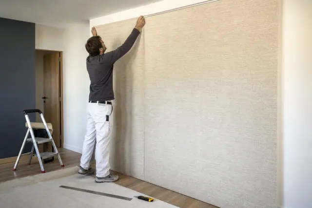 Poseur tend un lé de tissu beige sur un mur préparé dans un salon.