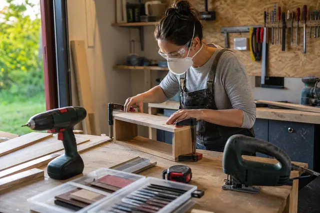 Bricoleuse équipée préparant une étagère avec visseuse et scie sur établi.