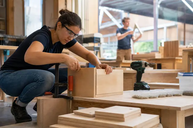 Débutante assemblant une caisse en bois dans un atelier collectif sécurisé.