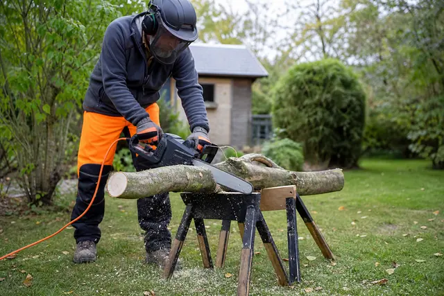 Jardinier équipé coupant une branche sur tréteaux avec une tronçonneuse électrique.