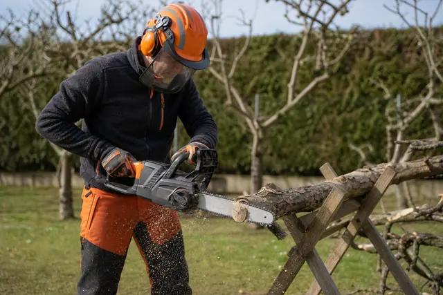 Jardinier équipé coupant une branche sur chevalet avec une tronçonneuse à batterie.